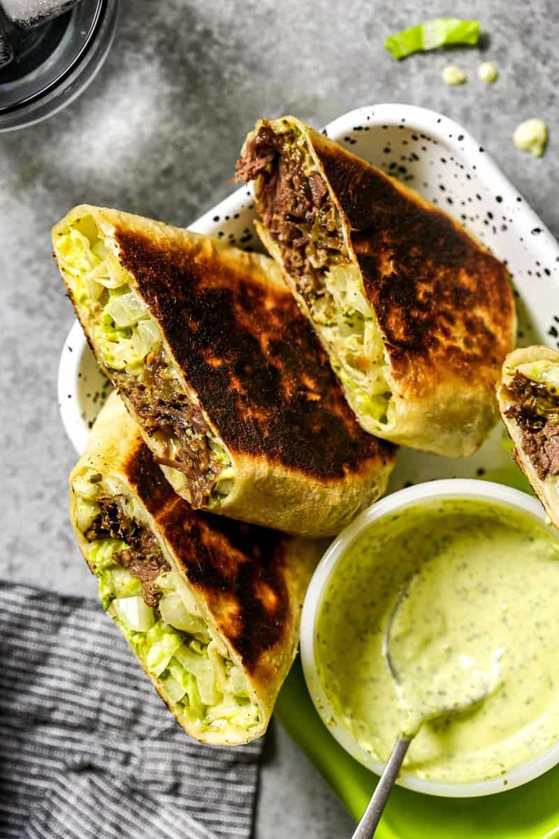 Three burrito halves arranged, cut-side up, on a white speckled metal platter with a small white bowl of chimichurri mayo set in it.