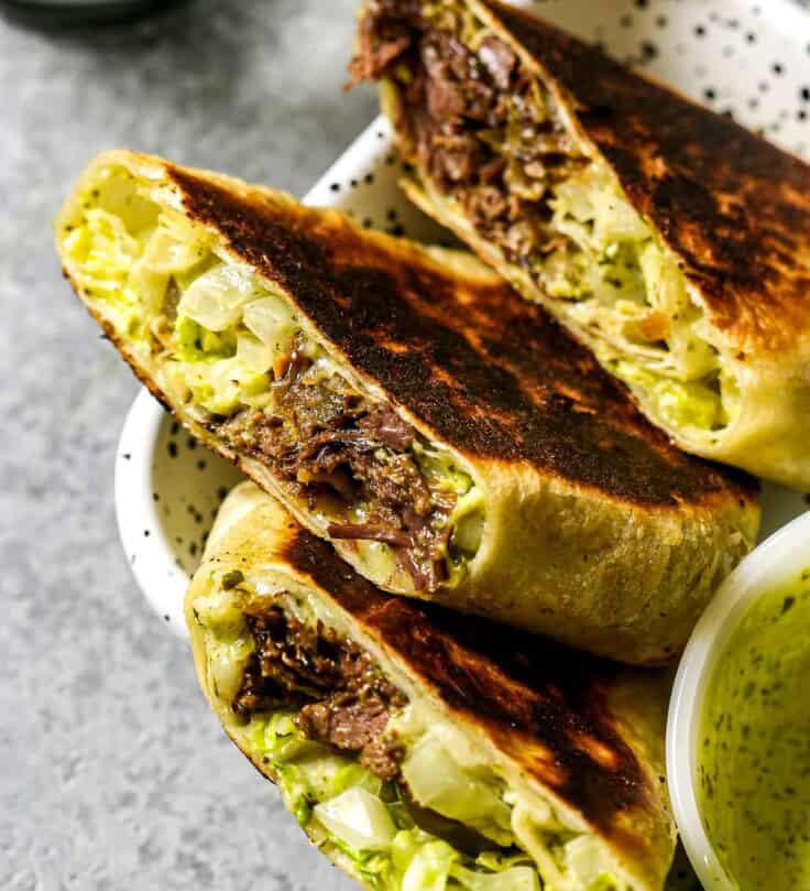 Three burrito halves arranged, cut-side up, on a white speckled metal platter with a small white bowl of chimichurri mayo set in it.