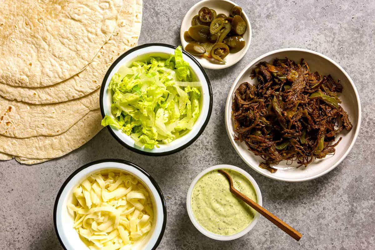 Flour tortillas, shredded cheese, shredded lettuce, pickled jalapenos, shredded beef, and chimichurri mayo set out on a counter.