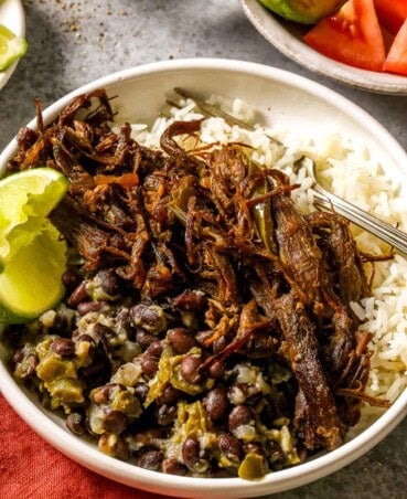 Shredded crispy beef (vaca frita) in a shallow white bowl with rice and seasoned beans. Two lime wedges also set in the bowl along with a silver fork. Chunks of tomato and avocado in a bowl set to the top right.