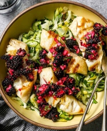 Large yellow serving bowl filled with a ribbon zucchini salad, four fillets of grilled sablefish and a blackberry relish, or salsa, spooned over top. Silver serving spoon set in bowl with a glass of ice water and a juiced lemon half set to the side.