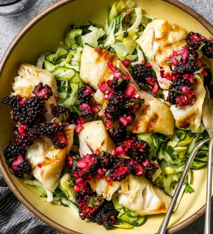 Large yellow serving bowl filled with a ribbon zucchini salad, four fillets of grilled sablefish and a blackberry relish, or salsa, spooned over top. Silver serving spoon set in bowl with a glass of ice water and a juiced lemon half set to the side.