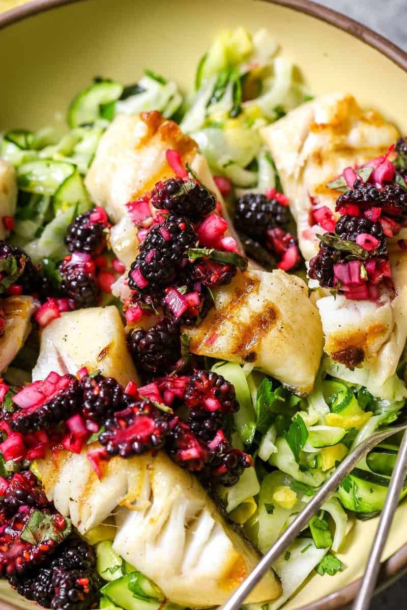 Large yellow serving bowl filled with a ribbon zucchini salad, four fillets of grilled sablefish and a blackberry relish, or salsa, spooned over top. Silver serving spoon set in bowl.