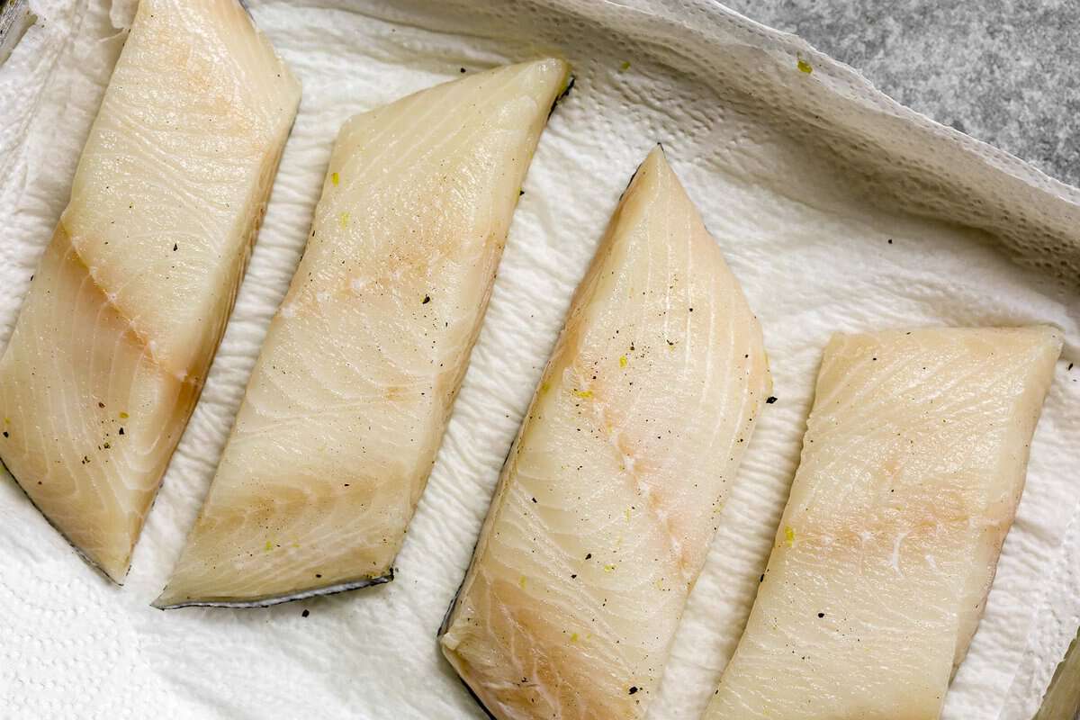 Sablefish fillets seasoned with lemon zest and black pepper on a paper towel lined sheet pan.