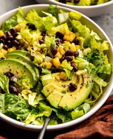 Chopped salad in a large white bowl with chopped romaine lettuce, black beans, charred corn, sliced avocado and a lime vinaigrette. a silver fork set in a salad.