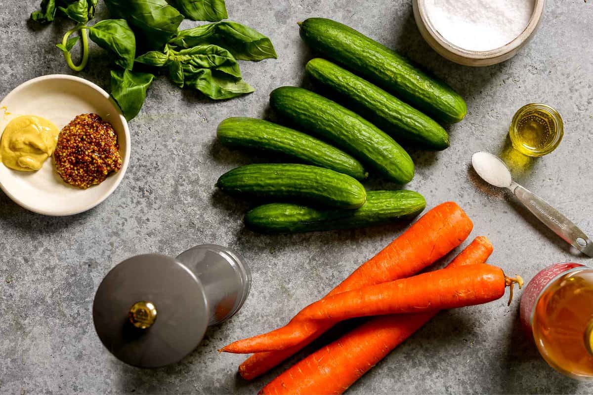 Persian cucumbers, three large carrots, olive oil, salt, pepper, sugar, vinegar, basil and mustard set out on a counter. 