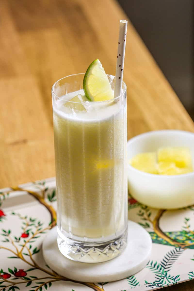 Frozen pina colada in a collins glass filled with ice, garnished with a lime wedge and a straw. Glass set on a patterned metal tray.