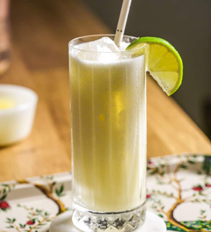 Frozen pina colada in a collins glass filled with ice, garnished with a lime wedge and a straw. Glass set on a patterned metal tray.