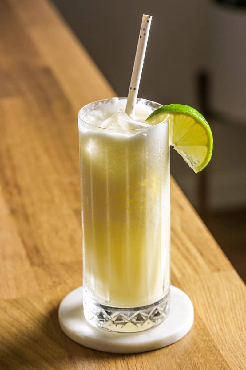 Frozen pina colada in a collins glass filled with ice, garnished with a lime wedge and a straw. Glass set on a white marble coaster on a wood table.