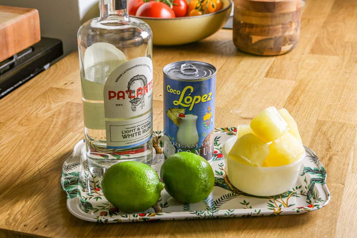 Rum, cream of coconut, limes and frozen pineapple juice set on a patterned metal tray.