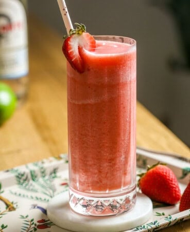 Collins glass filled with strawberry piña colada with a strawberry as a garnish and straw. Set on a patterned tray with fresh strawberries to the side.