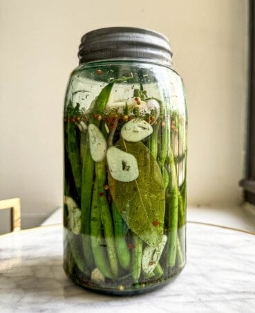 Green beans, sliced garlic and herbs in a tall blue-hued Ball jar filled with brine.