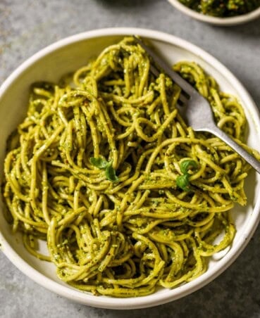 Pesto-coated pasta in a shallow white bowl with a silver fork set in it and tiny basil leaves scattered over top.