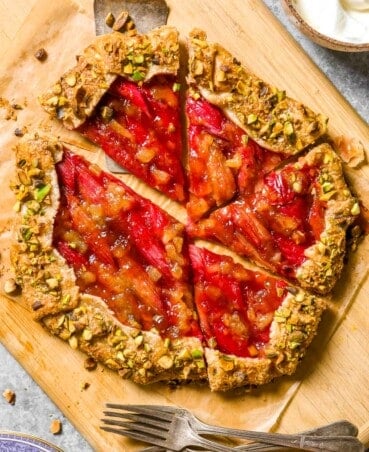 Rhubarb galette with toasted pistachios on the crust set on a wood cutting board lined with parchment paper. Silver forks, a bowl of whipped cream and serving plates set to the side.