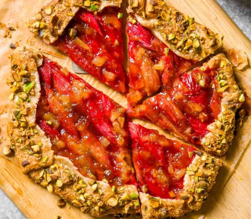 Rhubarb galette with toasted pistachios on the crust set on a wood cutting board lined with parchment paper. Silver forks, a bowl of whipped cream and serving plates set to the side.