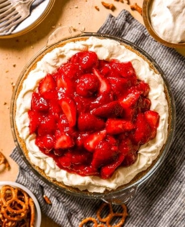 Pie plate lined with pretzel crust then filled with whipped cream cheese filling and topped with fresh strawberry filling set on a blue napkin with a bowl of pretzels and dessert plates with forks set off to the side.