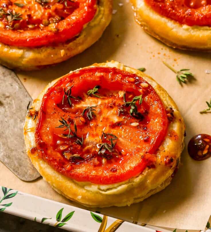 Three tomato puff pastry tarts on a parchment-lined serving platter with a silver serving spatula.