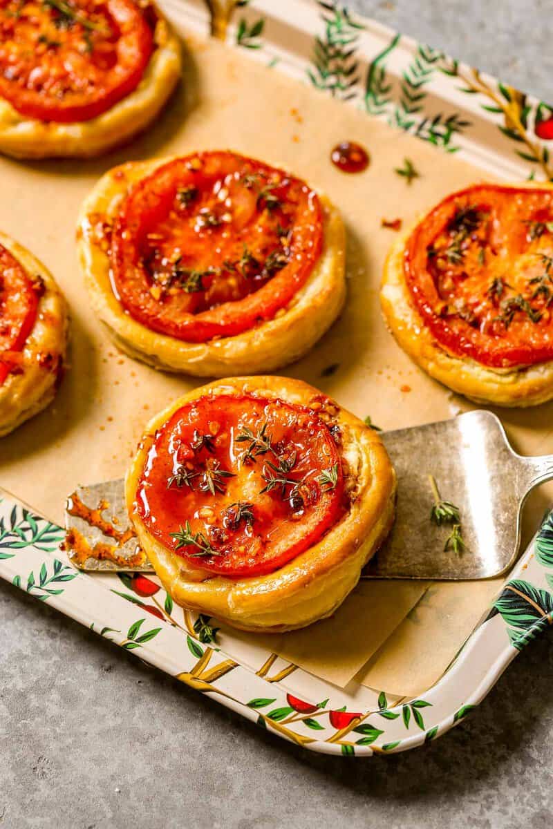 Five tomato puff pastry tarts on a parchment-lined serving platter with a silver serving spatula.