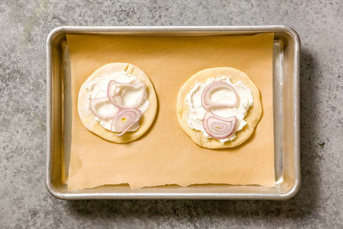 Two puff pastry rounds topped with goat cheese and slices of shallot on a parchment-lined baking sheet.