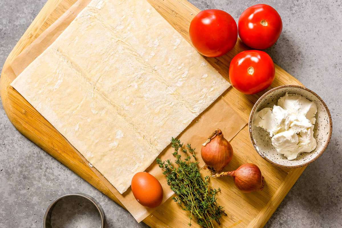 Wood cutting board topped with a sheet of puff pastry, three tomatoes, thyme, an egg, two shallots, goat cheese and a round cookie cutter.