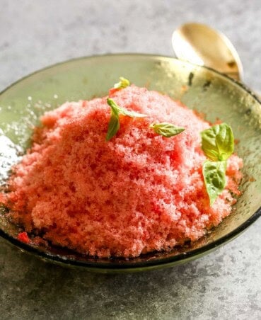 Shaved watermelon ice in a glass green bowl topped with fresh basil. Gold spoon set off to the side.