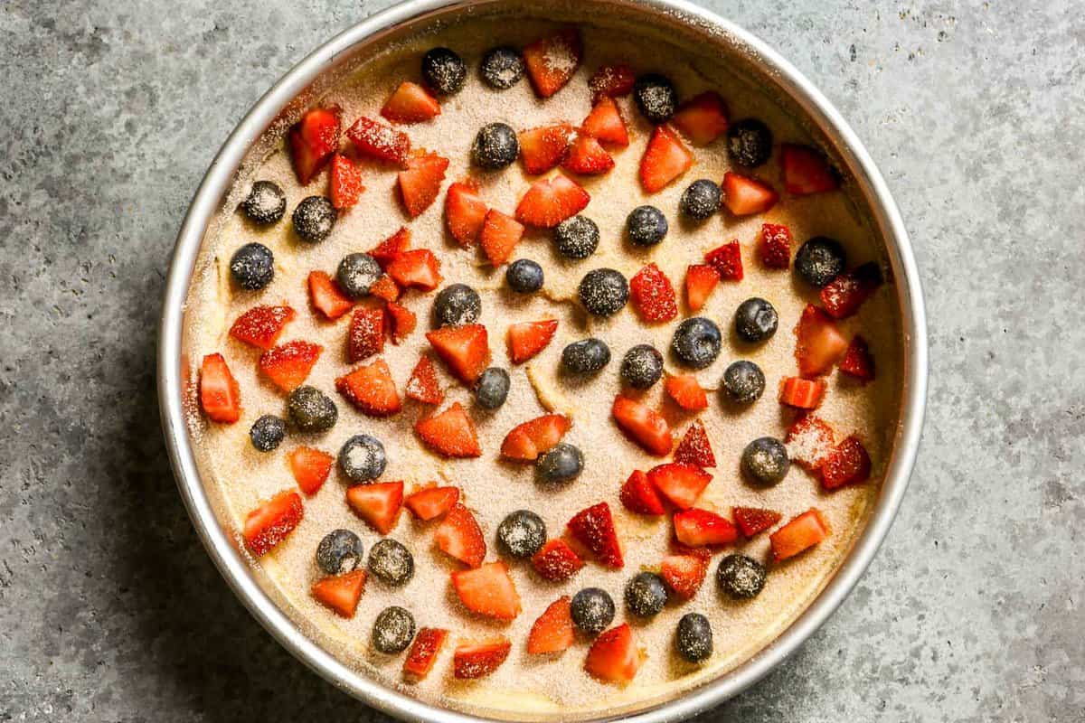 Round cake pan filled with cake batter and topped with diced strawberries, blueberries and cinnamon sugar.