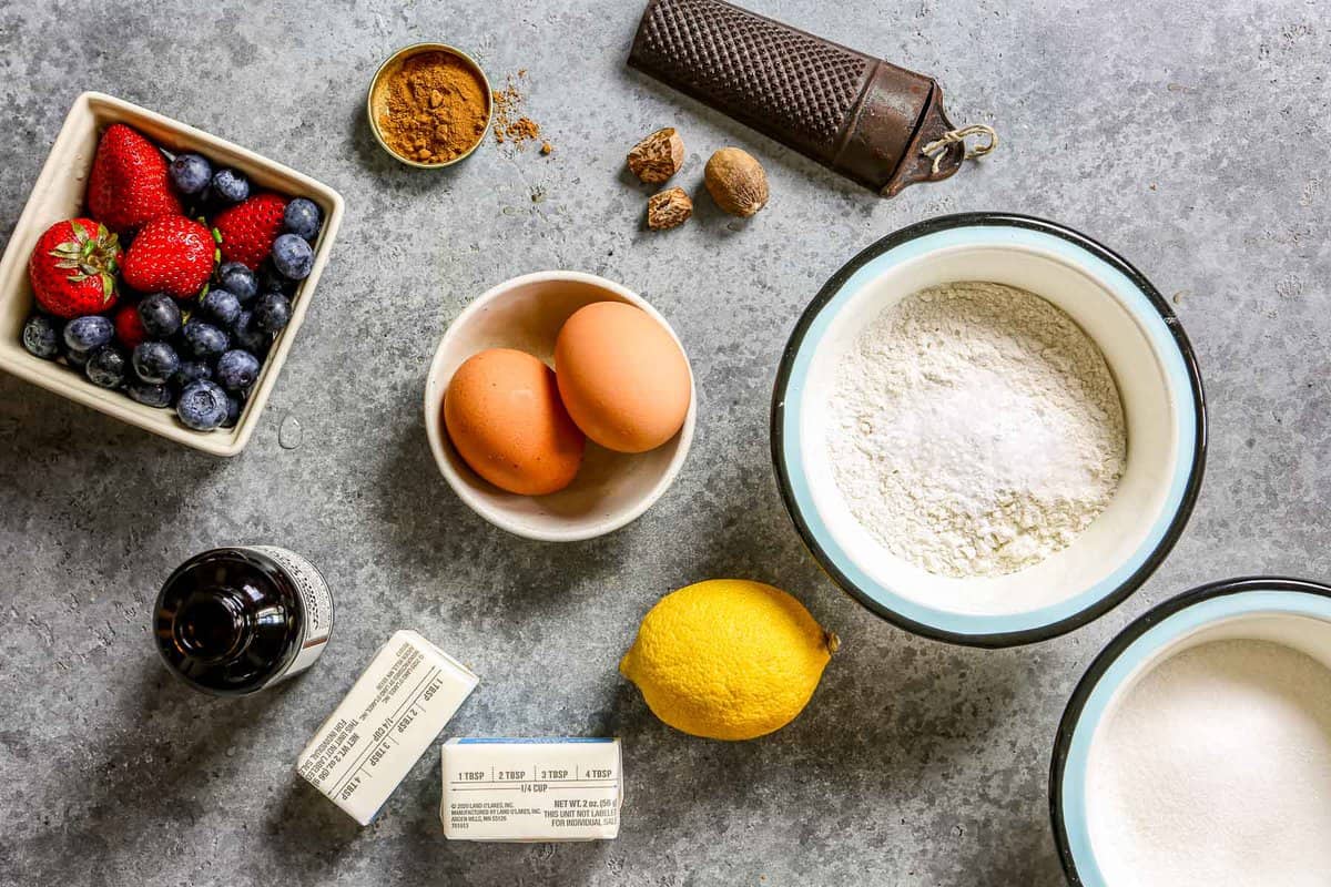 Fresh blueberries, strawberries, cinnamon, sugar, butter, nutmeg, lemon, flour and baking powder set out on a counter.