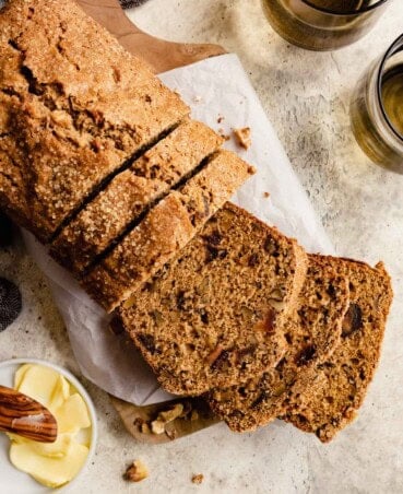 Date and nut bread set on a piece of parchment paper with a few slices cut from it. Butter and a glass of water set around.