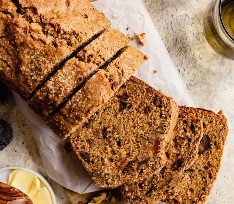 Date and nut bread set on a piece of parchment paper with a few slices cut from it. Butter and a glass of water set around.