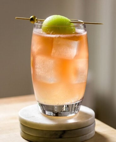 Honey deuce cocktail, light pink-orange in color, in a short water glass filled with ice, garnished with a honeydew melon ball and set on a stack of white marble coasters.