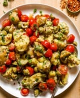 Torn chunks of potato tossed with halved cherry tomatoes, olives, fresh oregano and basil on a large white platter. Red pepper flakes and herbs set off to the side on wood countertop.