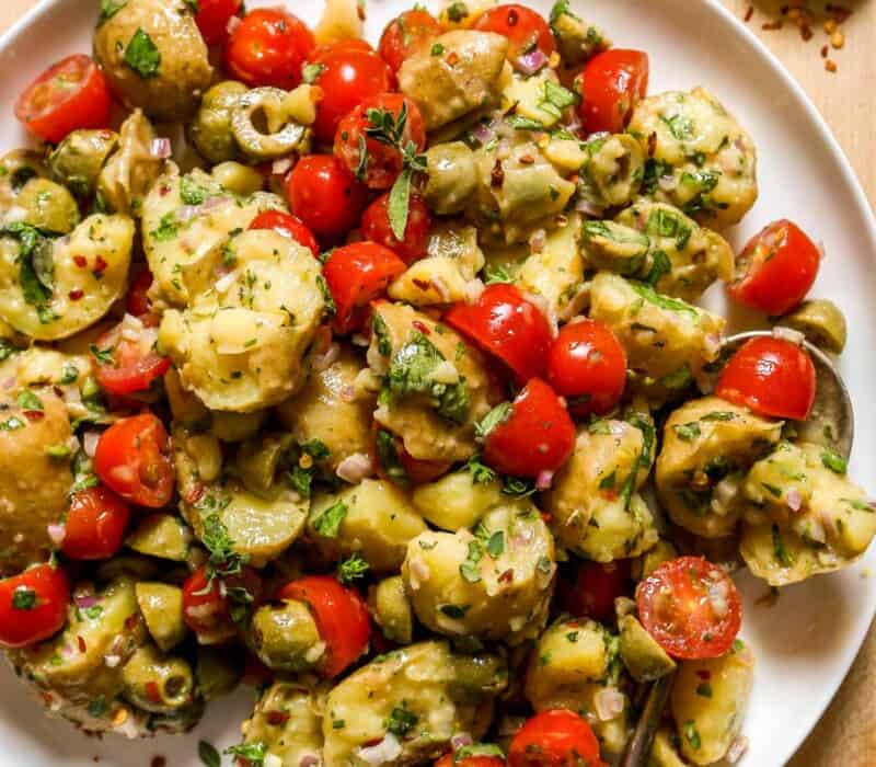 Torn chunks of potato tossed with halved cherry tomatoes, olives, fresh oregano and basil on a large white platter. Red pepper flakes and herbs set off to the side on wood countertop.