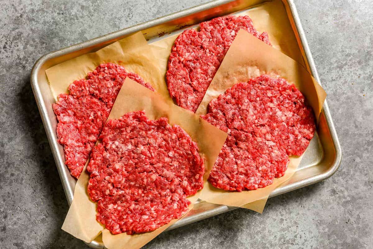 Ground beef smashed into thin patties and layered between parchment paper piled onto a baking sheet.