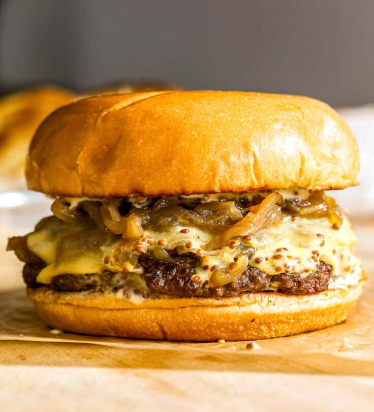 Smash burger topped with caramelized onions, cheese and sauce set on a piece of brown parchment paper on a wood cutting board.