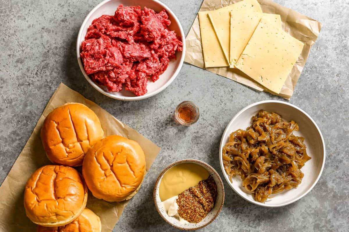 Ground beef, brioche burger buns, a bowl with yellow mustard, whole grain mustard and mayo in it, vinegar, 4 slices of deli cheese and caramelized onions set out on a counter.