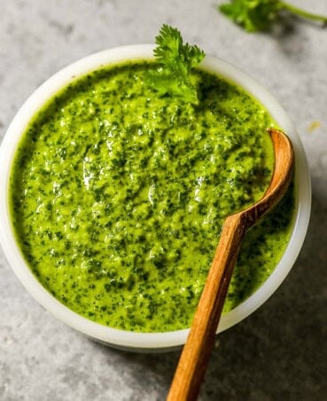 Vibrant green chimichurri sauce in a small white bowl with a small wooden spoon set in it. Fresh cilantro scattered around it on the counter.