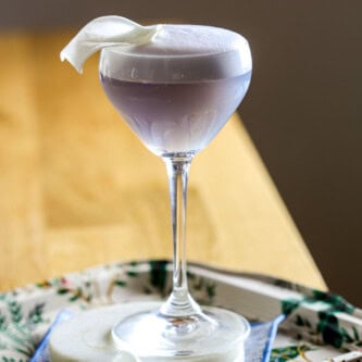 Purple Empress gin cocktail in a coupe glass topped with a white foam top and flower petal. Set on a stack of marble coasters.