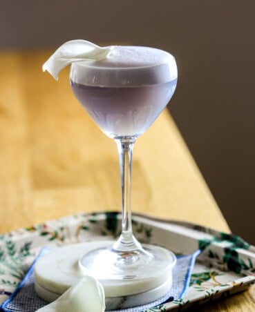 Purple Empress gin cocktail in a coupe glass topped with a white foam top and flower petal. Set on a stack of marble coasters.