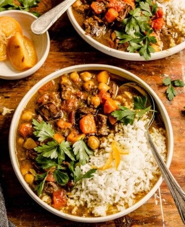 Instant pot lamb stew with chunks of carrots and chickpeas in a rich, thick broth in a shallow white bowl with white rice, parsley and sliced preserved lemon on top.