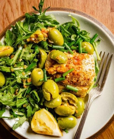 Golden brown cod fillets on a plate with fresh arugula, smashed olives, butter sauce and lemon wedge.