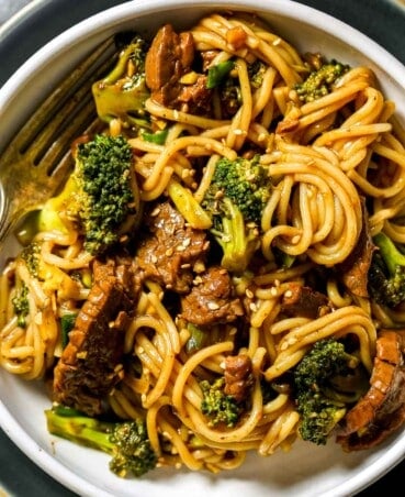 Beef and broccoli noodles in a shallow white bowl set on a blue plate with a silver fork set in the bowl. Sesame seeds in a small wooden bowl set off to the side.