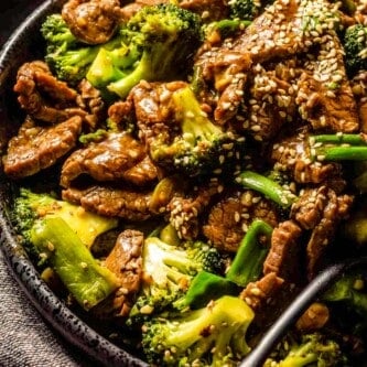 Stir-fried beef and broccoli in a large black serving bowl with sesame seeds scattered over top and a silver serving spoon set in it.