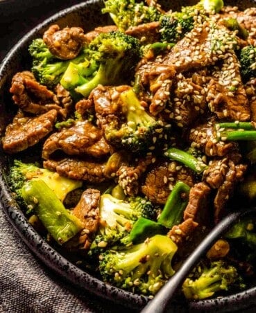 Stir-fried beef and broccoli in a large black serving bowl with sesame seeds scattered over top and a silver serving spoon set in it.