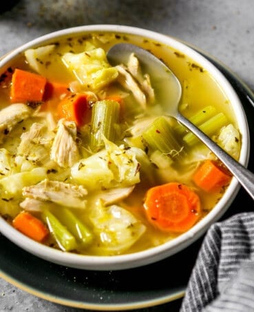 Chunks of cabbage, carrots and celery in a chicken and cabbage soup in a shallow white bowl with a silver spoon set in it.