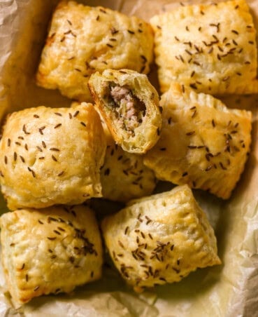 Baked ham and cheese puff pastry bites with caraway seeds on top set on in a parchment-lined metal serving platter. Top puff pastry bite with a bite taken out.