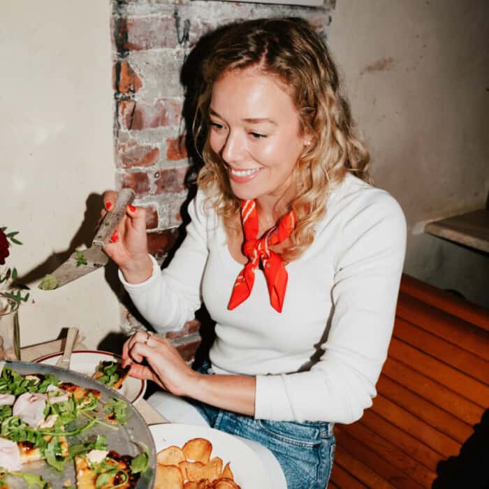 Lauren Grant, in a white long sleeve shirt with a red scarf around her neck, picking up a piece of pizza with a spatula.