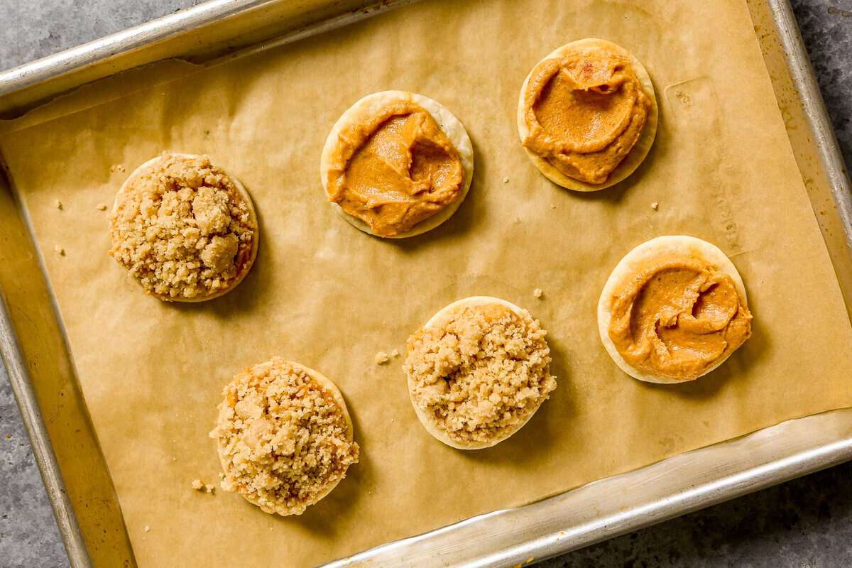 Six pumpkin pie cookies set on a parchment-lined baking sheet. Three with filling spread on top of the pie crust and three with streusel stuck to filling on crust.