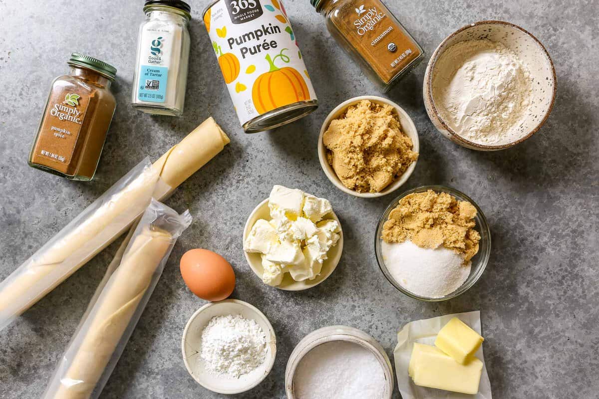 jars of pumpkin pie spice, cinnamon and cream of tartar set out on a counter with a can of pumpkin puree, premade pie crust dough, an egg, butter, brown sugar, granulated sugar, flour, salt, and cornstarch.