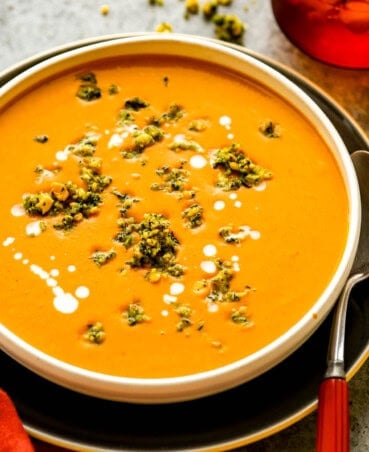 Creamy sweet potato carrot soup in a shallow white bowl set on a plate with crumbly herb topping and coconut milk drizzled over the soup.