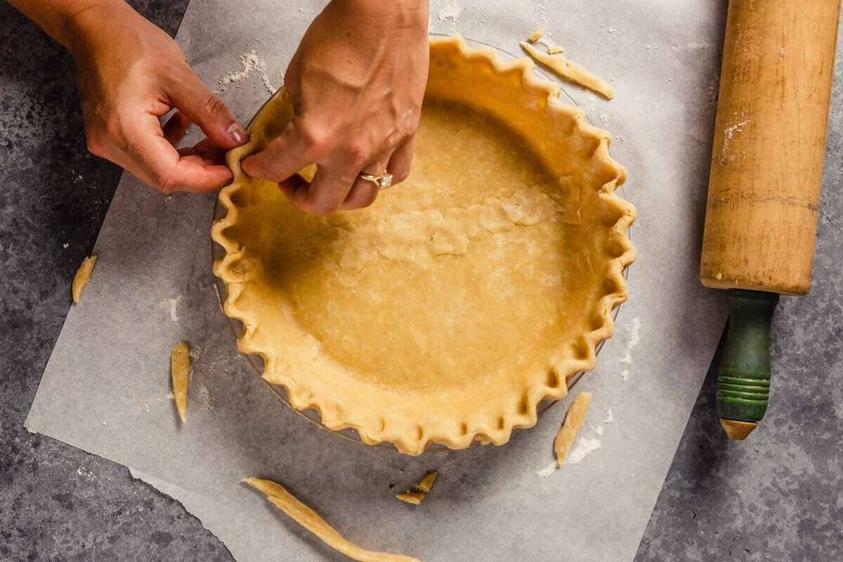 A set of hands crimping the edges of pie dough in a clear glass pie plate.